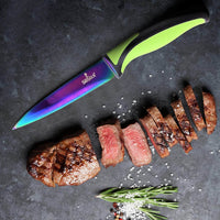 Rainbow titanium coated steak knife with green handle, beside sliced steak and fresh herbs on a dark countertop.